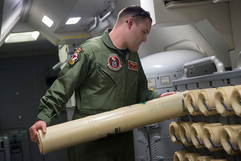 A US naval officer loads a sonobuoy onto a rack for use in the search for Flight MH370. He wears an army green jumpsuit and has a buzz-cut hair style. The sonobuoy he is loading is cylindrical and beige in colour. He appears to be on a aeroplane. 