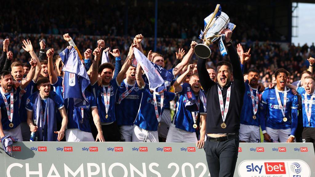 John Mousinho lifts the League One trophy above his head while the Portsmouth players raise their arms in celebration in the background.