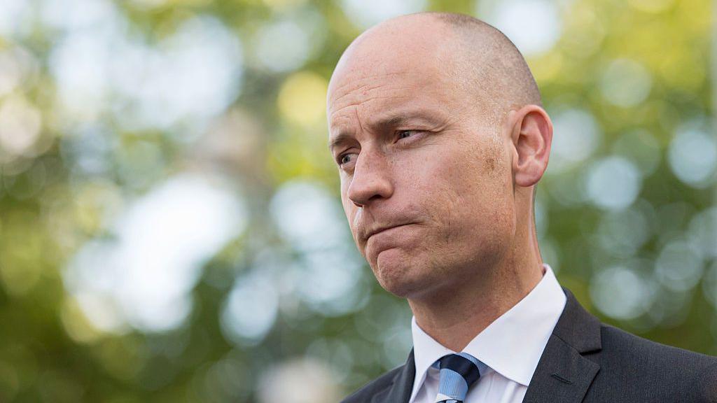 Stephen Kinnock wearing a white collared shirt, a black suit and a black and blue striped tie. He had a bald head and is clean shaven. He is looking towards the left of the camera with a thoughtful expression. There are trees in the background, out of focus.