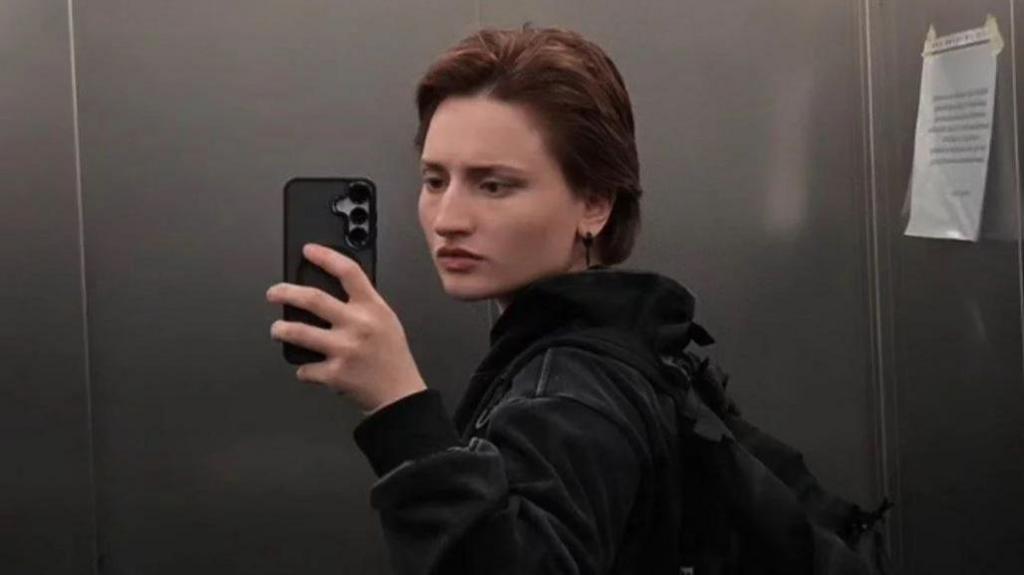Aishat taking a mirror selfie inside an elevator. She is wearing a dark hoodie and carrying a black backpack