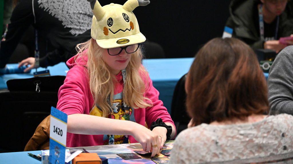 A young Pokémon fan sits across a table from an opponent during a match of the trading card game. Wearing a yellow baseball cap with Pikachu ears and a bright pink hoody with a colourful Pikachu print on it, she looks thoughtful as she considers her next move. 