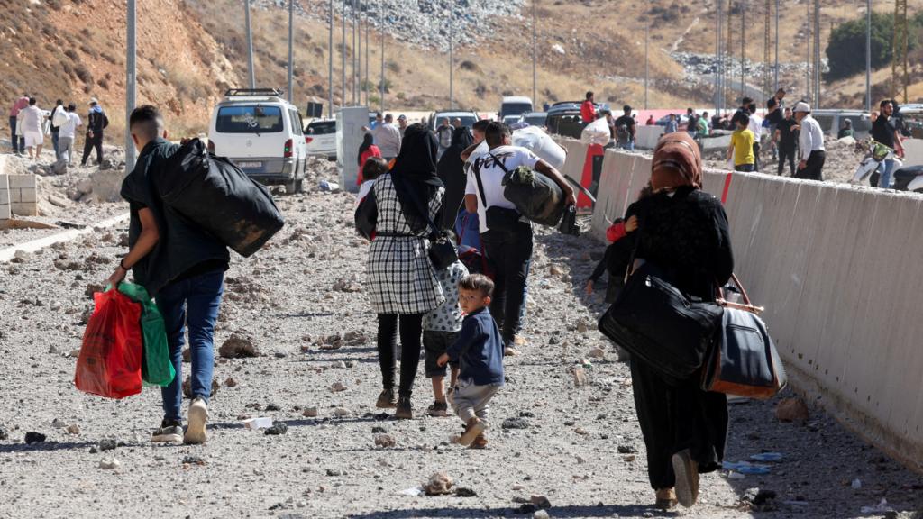 People carry their belongings while walking on the rubble, after an Israeli strike, as they flee Lebanon at Masnaa border