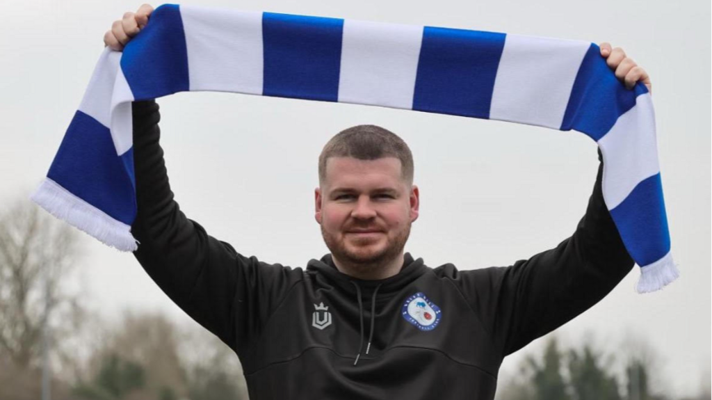 Aaron Hunt holds up a blue and black football scarf while wearing a black hoodie bearing the logo of Daisy Hill FC. He is stood outside with trees seen in the background. 