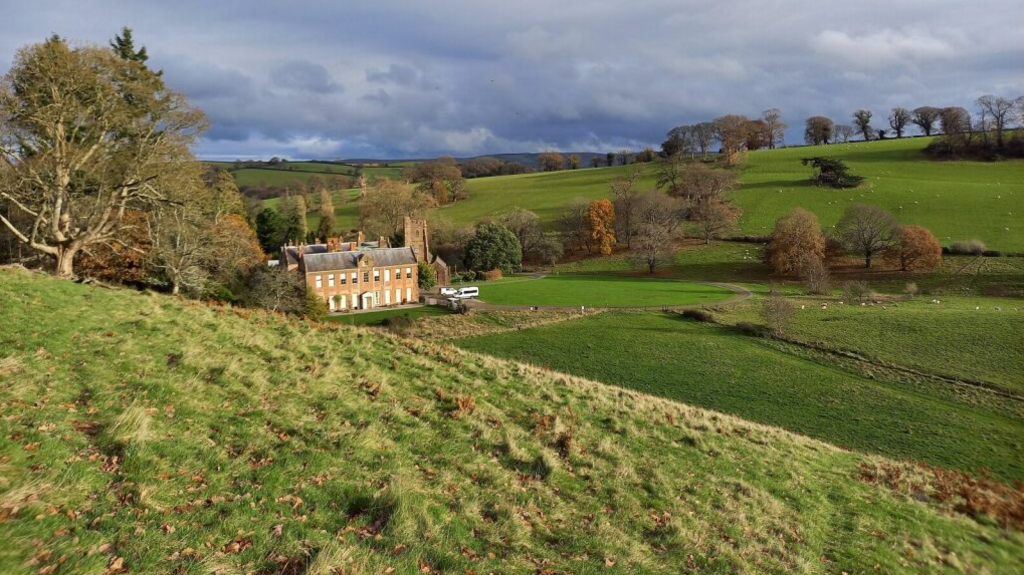 The image shows Nettlecombe Court, a grand house, nestled between green fields and trees.  