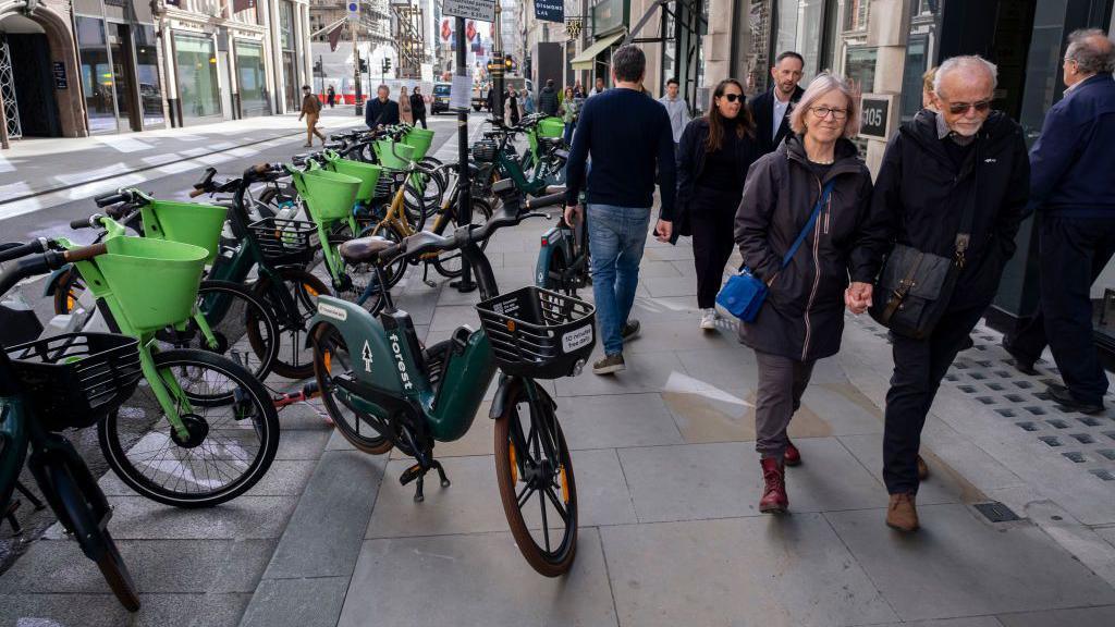 People walk past a row of parked rental bikes, including green and dark green Forest e-bikes, on a busy city street lined with shops and historic buildings.