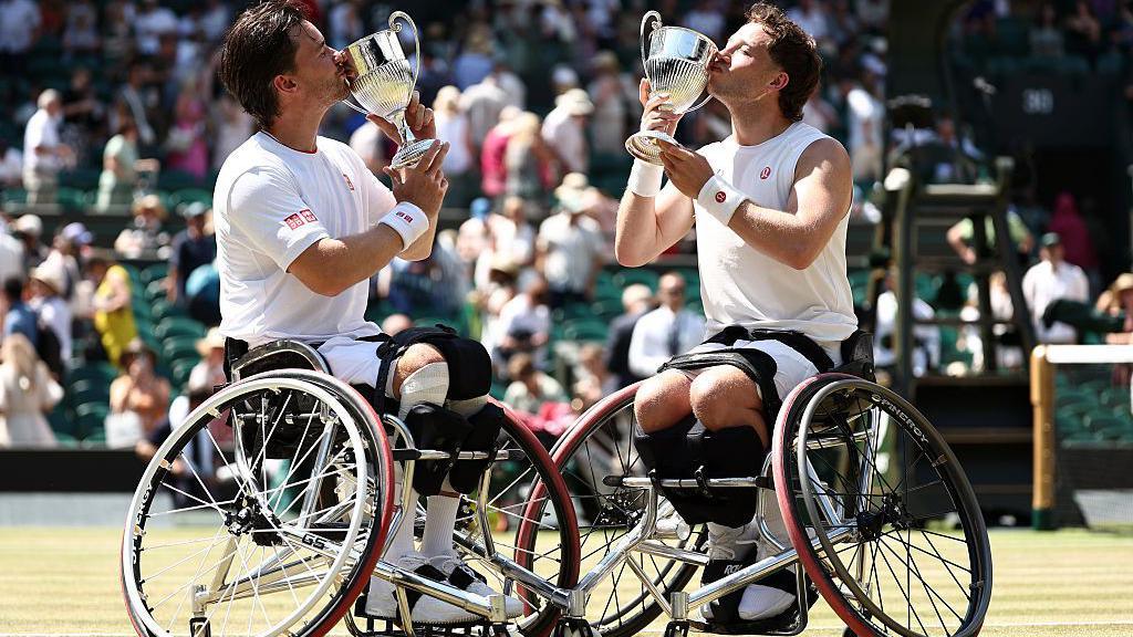 Gordon Reid and Alfie Hewett of Great Britain kiss their trophies following their win. They are both wearing white shorts and tshirts and are holding up their trophies to kiss them.