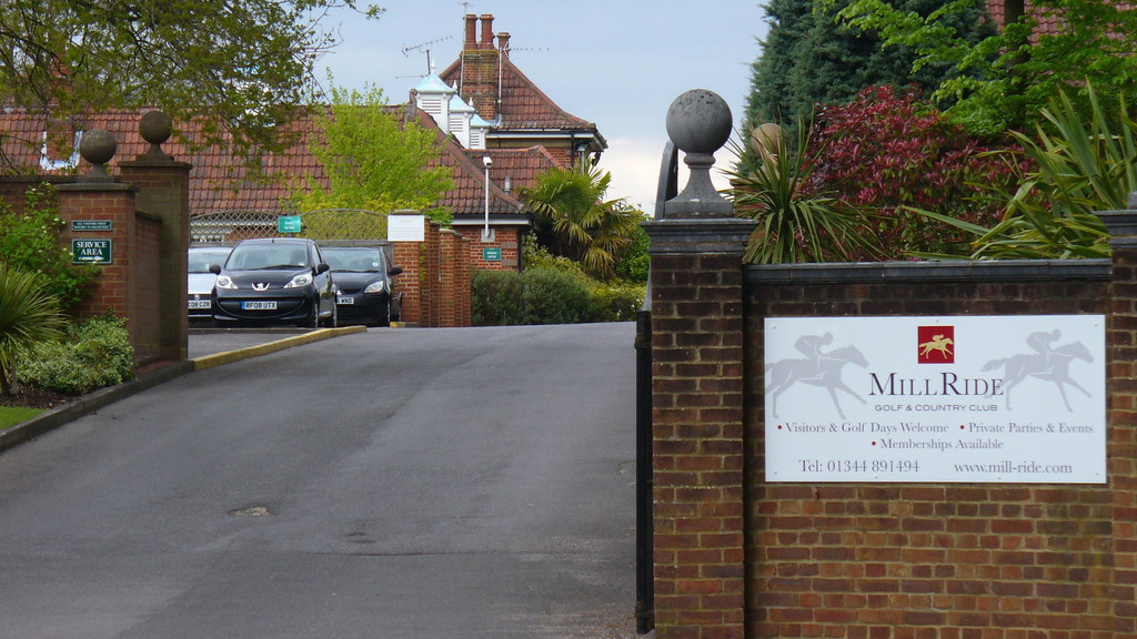 The entrance to Mill Ride golf and country club, which has a sign on a wall outside it and other buildings further up a road.