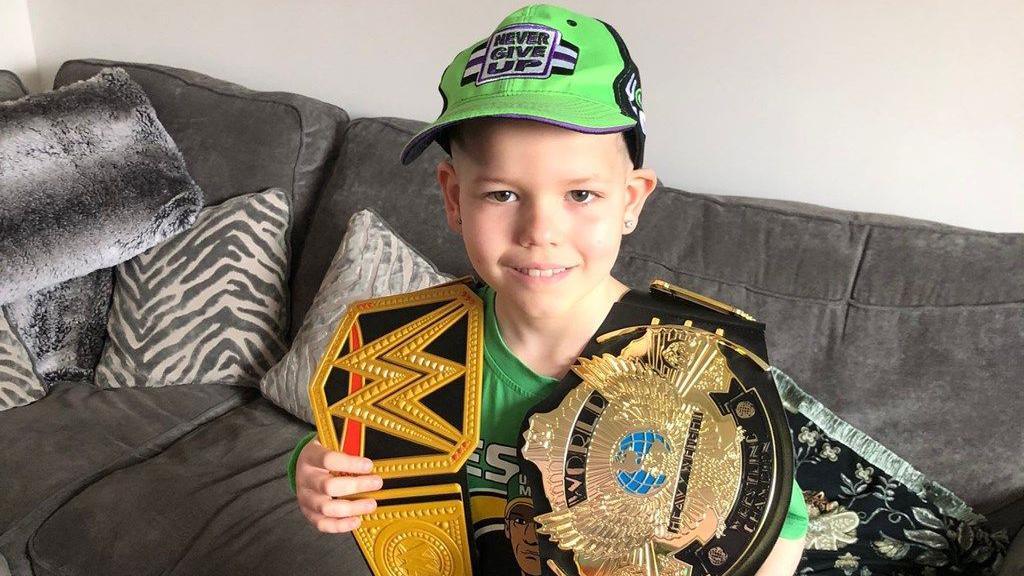 A picture of an eight-year-old boy in a green cap and tshirt holding two golden kicking-boxing belts and standing in front of a grey sofa