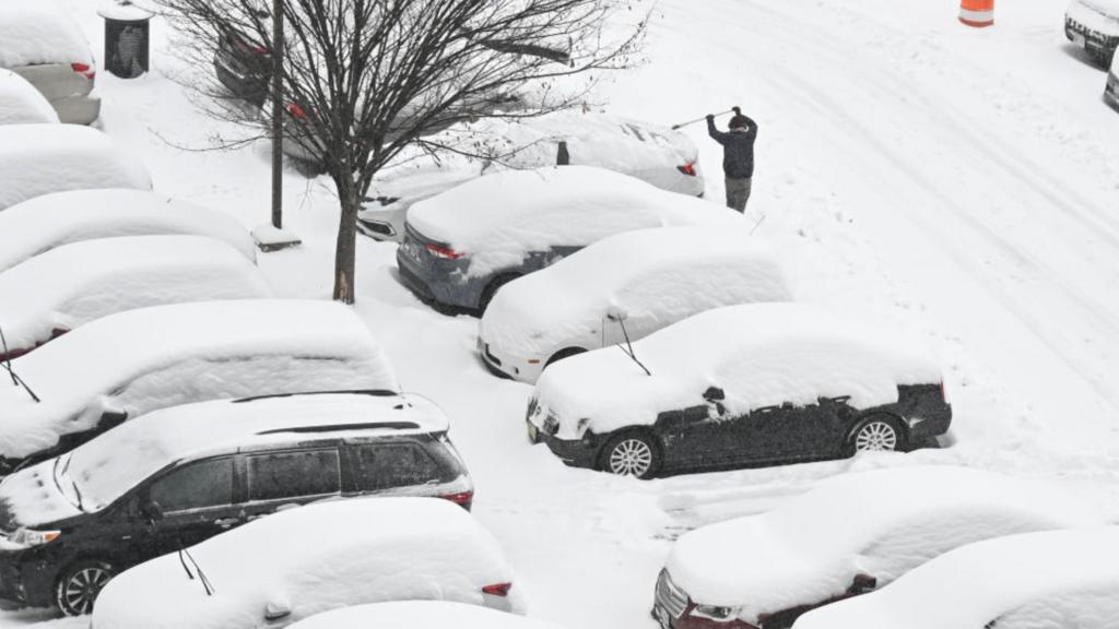 A person shovels snow from a car, with other cars parked nearby covered in a thick layer of snow