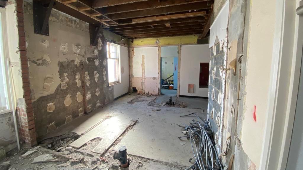 Interior of a room which has been stripped out. There is exposed plasterwork on all the walls, debris - including a bundle of what appears to be thin pipes or thick wires - and an exposed ceiling beams