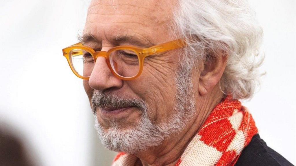 A man with white/grey hair looks away from the camera smiling. He is wearing orange glasses and a white and red scarf.