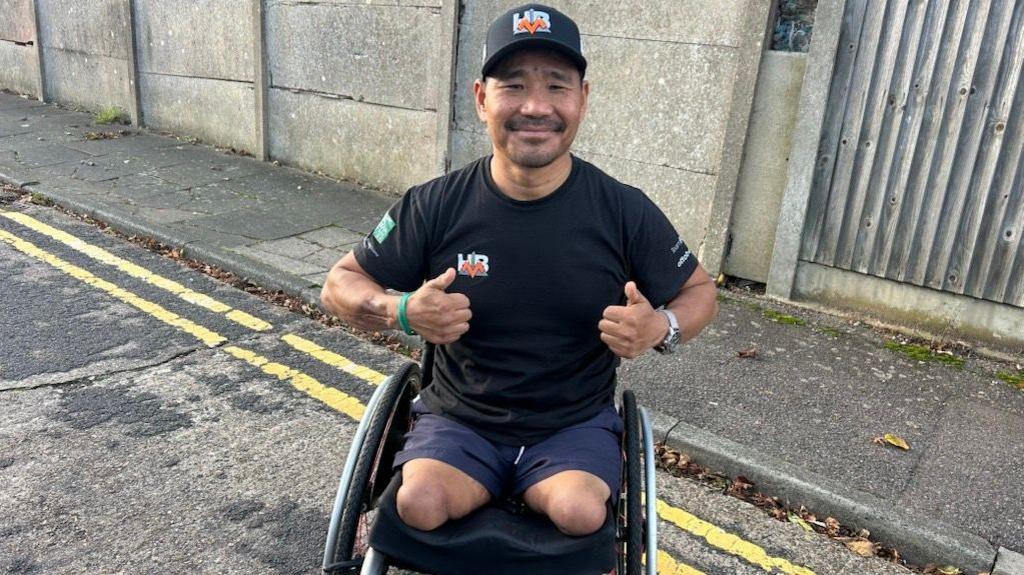 Hari Budha Magar sat in his wheelchair smiling and wearing a cap in a street with a wall behind him 