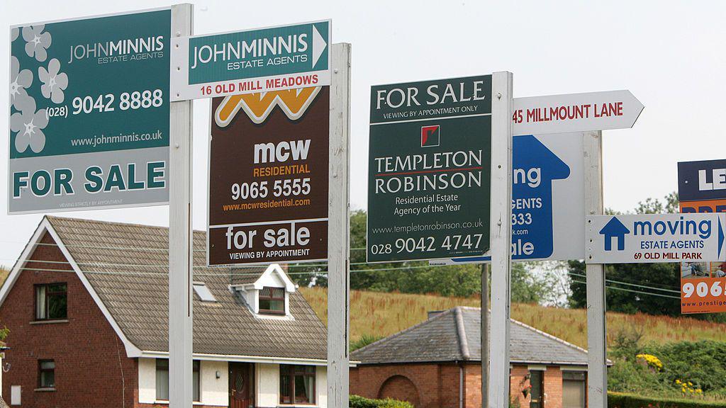 A row of 'For Sale' signs in front of two houses.