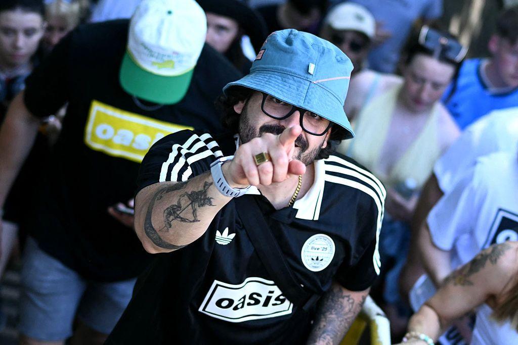 A man in a blue bucket hat and a black Oasis T-shirt points at the camera