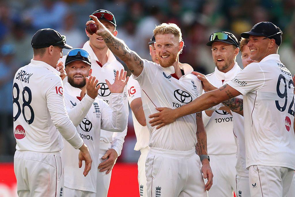 England cricket players in white uniforms gather on the field, celebrating a wicket. They wear caps and sunglasses, with visible sponsor logos on their shirts, and appear to be in a stadium under bright sunlight.