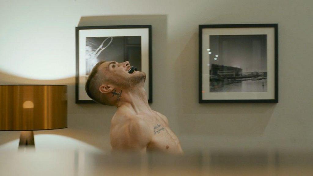 Pete looking up at the ceiling. He is shirtless and has a mouth guard in his mouth, seemingly doing his boxing training. Behind him are two pictures hanging on the wall, and a bronze lamp. 