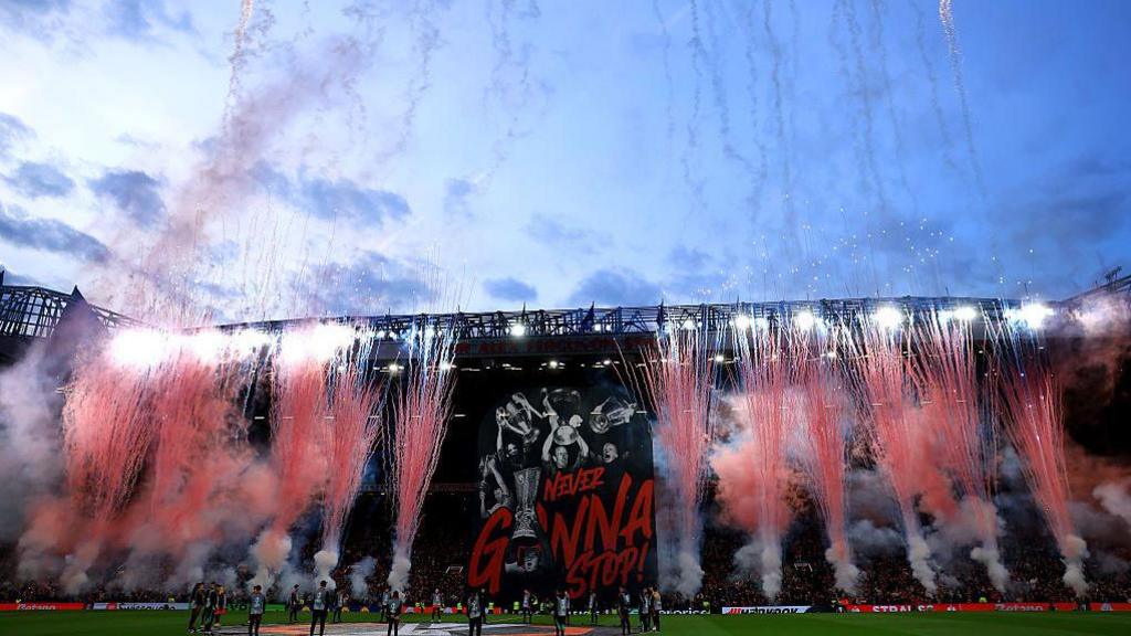 Manchester United's tifo prior to their Europa League fixture against Lyon. Pink fireworks are in front of a tifo with red writing on a black background.