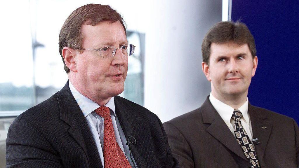An older photograph of two men. The older man on the left has short brown hair and is wearing small metal framed glasses. He is wearing a dark suit, light blue shirt and red patterned tie. The younger man on his left is looking at the older man with a raised eyebrow and slight grin. He he has black hair and is wearing a dark suit, white shirt and black patterned tie.