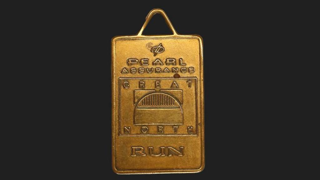 A gold rectangular shaped medal that reads 'Pearl Assurance' at the top of it and then in a square reads great north with a bridge, and underneath it reads run.