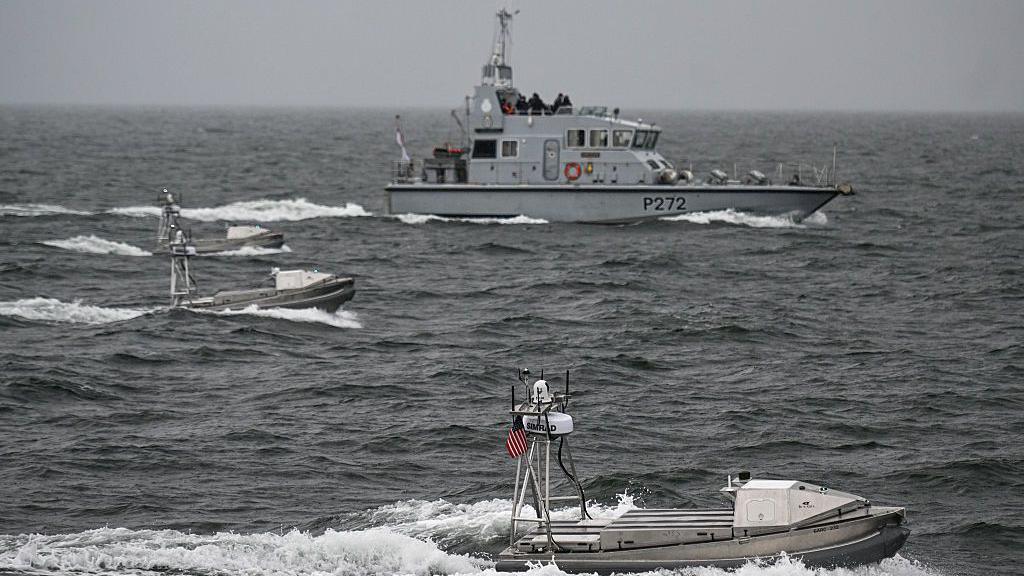 Three small unmanned boats sail through choppy grey seas in front of a larger grey navy boat 