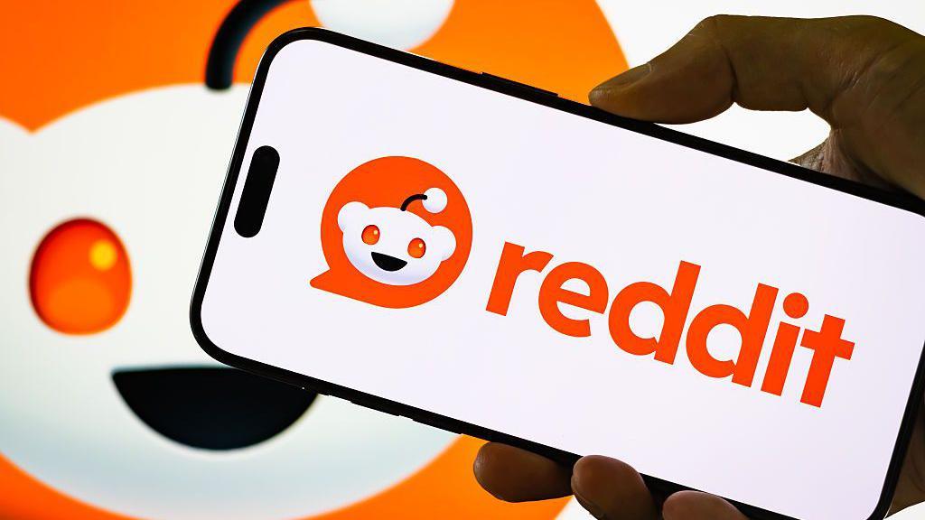 A hand holding a mobile phone with the reddit logo with the reddit character in the background