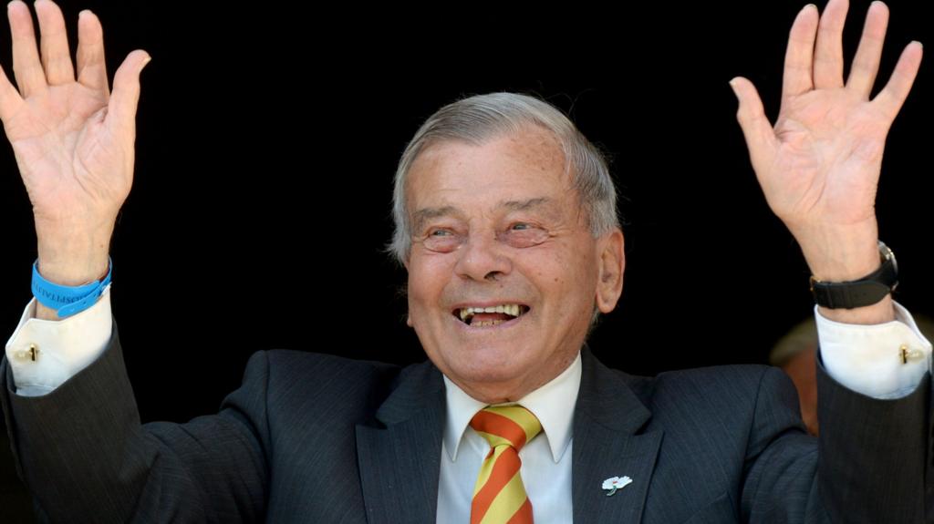 A man in a suit smiles and raises his hands