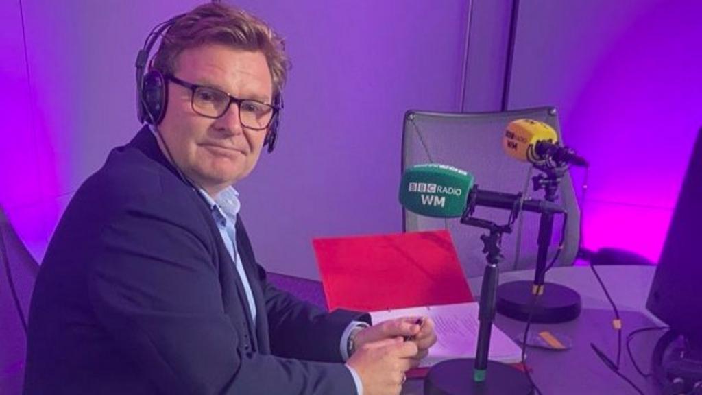 A man with short light brown hair wearing a navy suit sits in a purple radio studio