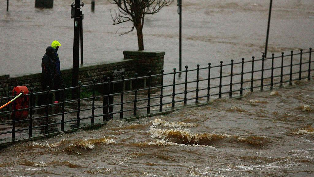 Two people on the Whitesands in Dumfries look at water lapping up from the River Nith across a nearby car park