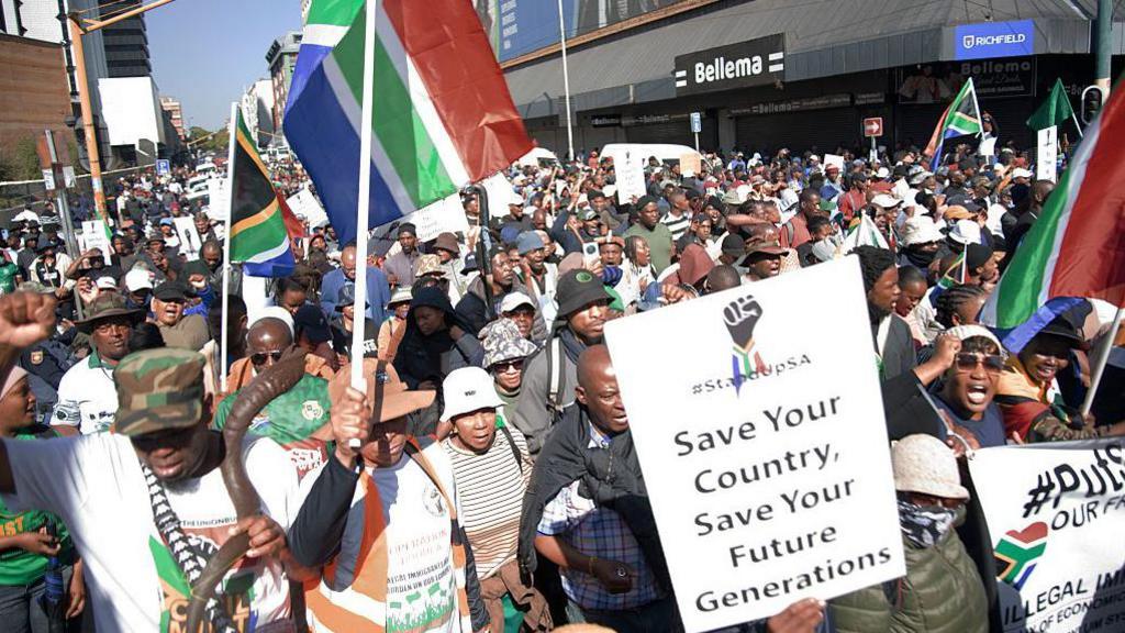 A crowd of people on a demonstration. Some are holding South African flags and at the front there is a placard that says "Save your country, save your future generations".