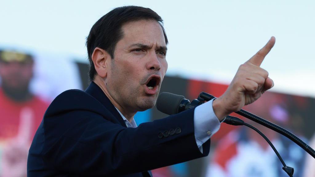 Marco Rubio, wearing a suit, gestures with the index finger of his right hand, as he speaks at a Trump rally