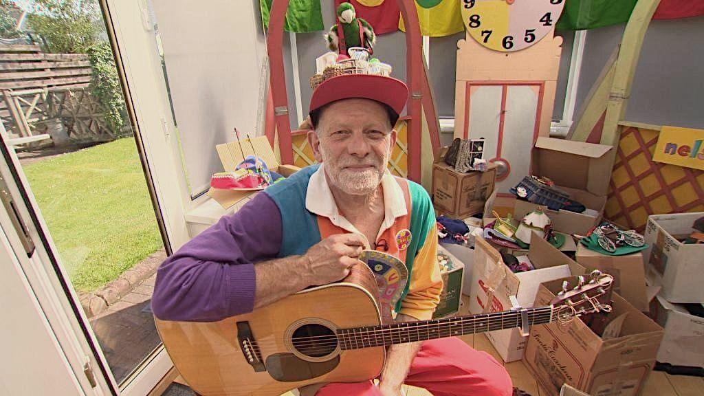 Donnie Dòtaman na shuidhe le giotàir air a' ghlùin, ad annasach air a cheann agus fiamh-ghàire air aodann. Tha e ann an seòmar làn bhogsaichean agus gleòc Dòtaman air a chùlaibh. Tha coltas ann gur e 'props' bho Dhòtaman a th' air a chùlaibh. 