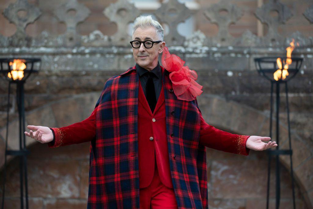 Alan Cumming in a tartan and bright red suit and shawl, arms open wide, smiling outside the castle in The Traitors