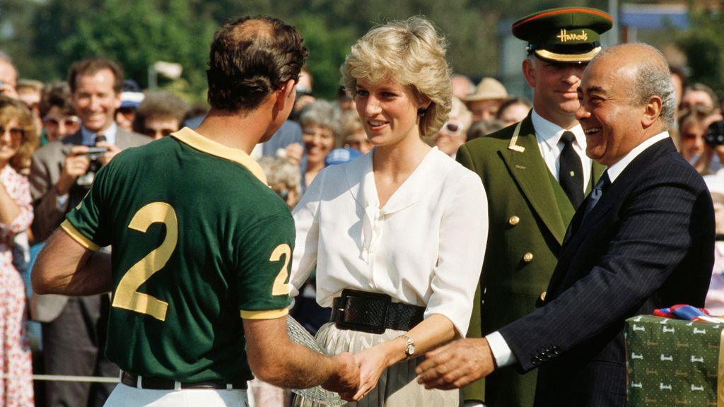 Princess Diana, pictured smiling in the 1980s, handing a glass trophy plate to Prince Charles, who has his back to the camera wearing a sports t shirt with a number two written on it. She has short blonde hair, a white blouse and chunky black belt. Mohamed Al Fayed is pictured at the right of the shot, smiling at them both, in the conversation, wearing a dark suit. A crowd of spectators are behind them. 