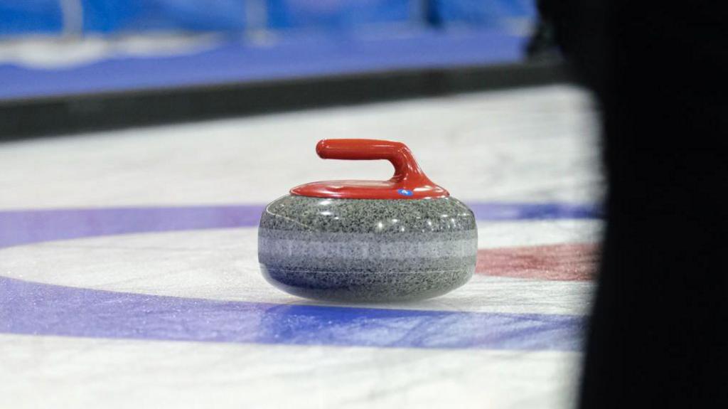 A curling stone with a red handle sitting on an ice rink