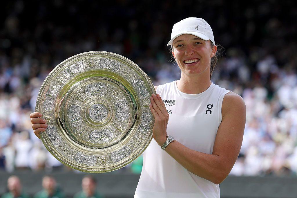 iga swiatek holding the ladies' single trophy while smiling in front of a crowd at centre court in wimbledon