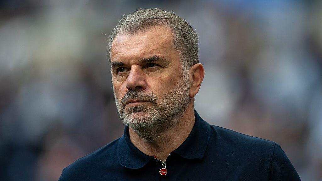 Close-up of Ange Postecoglou. He has short grey hair, brushed away from his face, and a grey beard. He is wearing a navy top which has a zip fastening at the collar.
