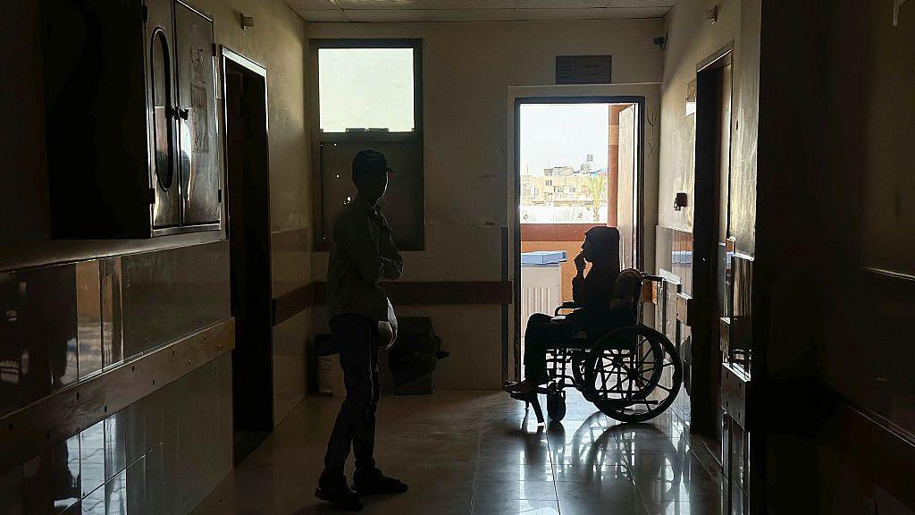 A person stands near a patient sitting in a wheelchair in a dark corridor during a power outage at Nasser hospital in Khan Younis, southern Gaza (20 June 2026)