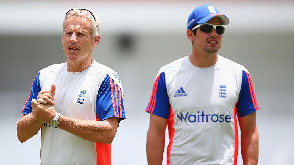 Peter Moores, to the left and Alastair Cook, to the right both in England training kit (mainly white with red and blue detailing). Looking at an angle away from each other. Moores has glasses up on his head. Cook wears sunglasses and a blue England cap.