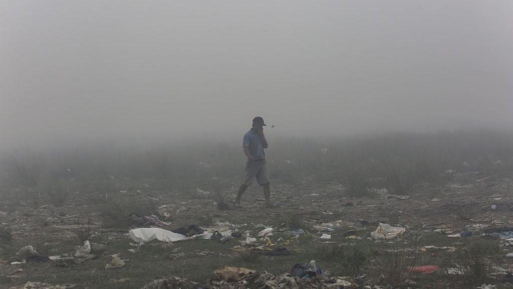A person walks through fog over rubbish-strewn ground while holding a hand over his mouth