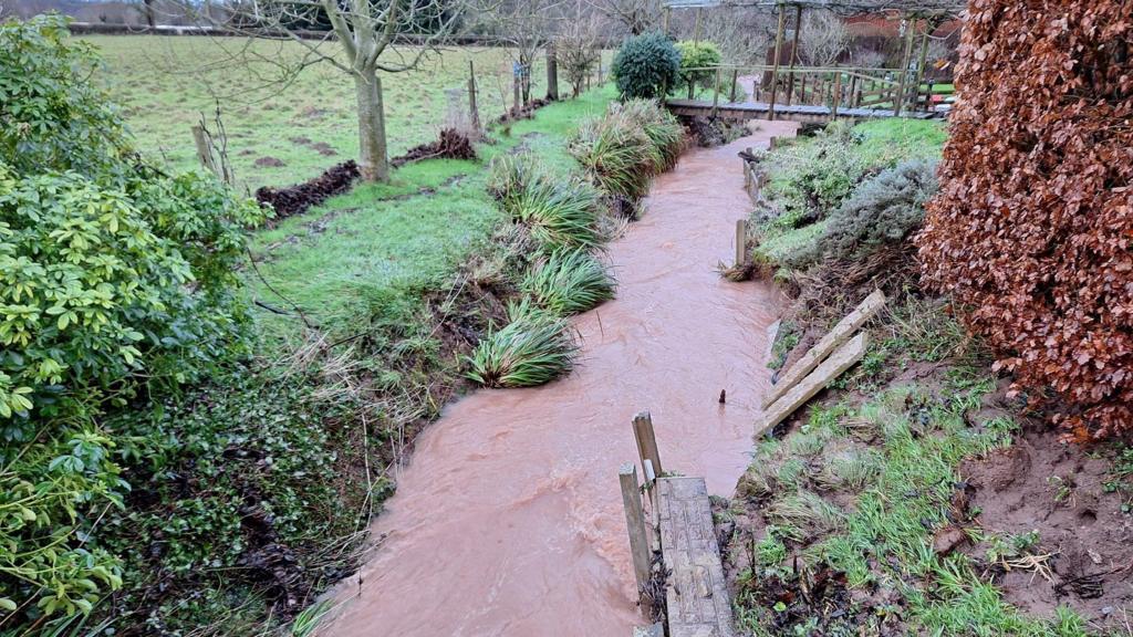 A swollen brook runs between fields and under a wooden bridge with trees visible in the distance.