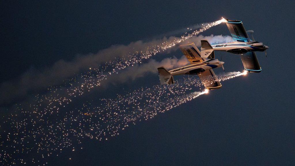 Two Firebirds planes fly upside down close to one another through a dark sky with pyrotechnic sparks coming from the tips of their wings.