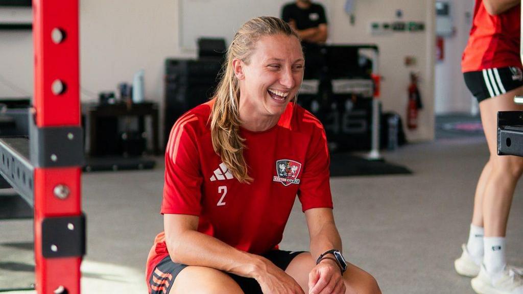 Bow Jackon wearing a red football shirt, hunched in a gym setting with a big smile on her face