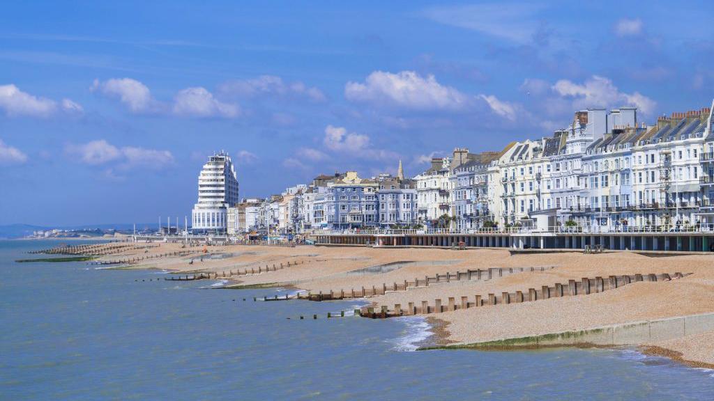 The promenade of an English seaside town. It is sunny. There is a shingle beach.