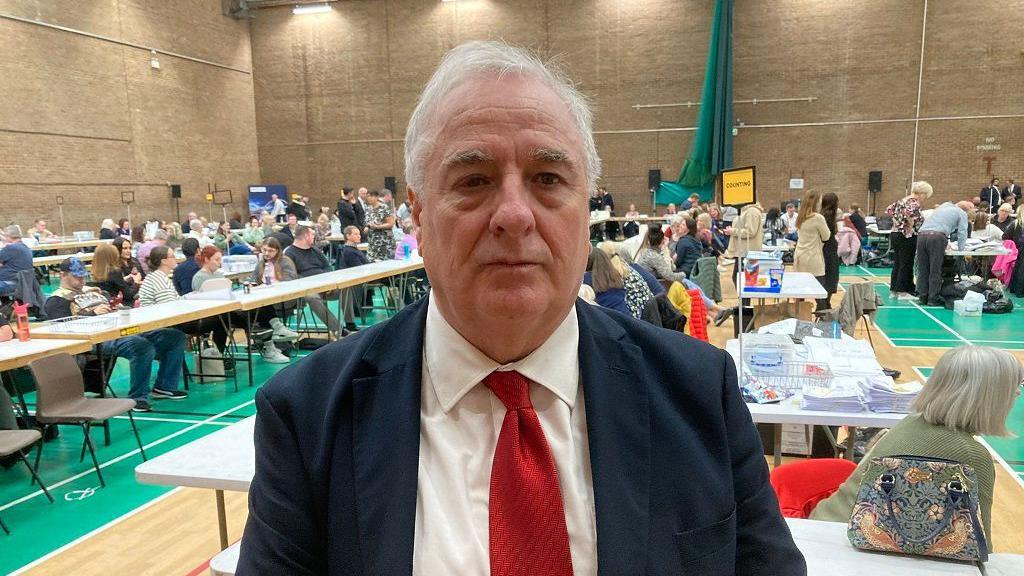 Head-and-shoulders image of Bob Cook attending an election count at Thornaby Pavilion. He has grey hair and is looking directly into the camera. He is wearing a navy suit, white shirt and red tie. Dozens of people are counting votes on tables behind him.