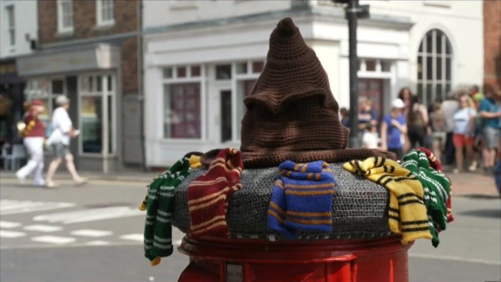 A knitted post box topper. It is a brown wizards hat, also known as the sorting hat. Around it are knitted colourful scarves of the Hogwarts houses colours - blue, red, yellow and green.