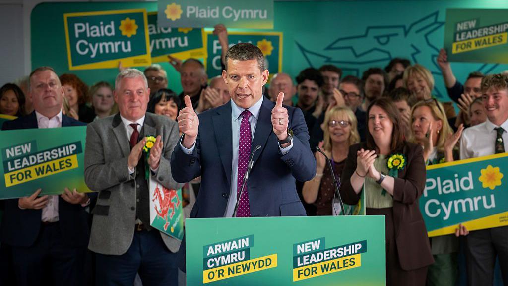 Politician Rhun ap Iorwerth gives a thumbs up at a Plaid Cymru event that says New Leadership for Wales.