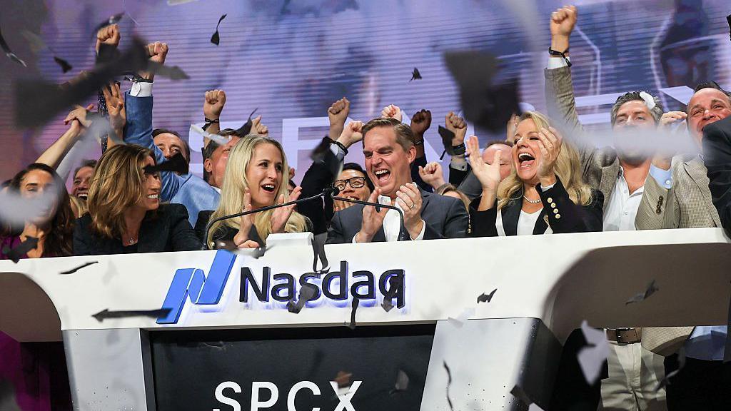 Senior people in SpaceX celebrate as confetti rains down in front of a sign saying Nasdaq