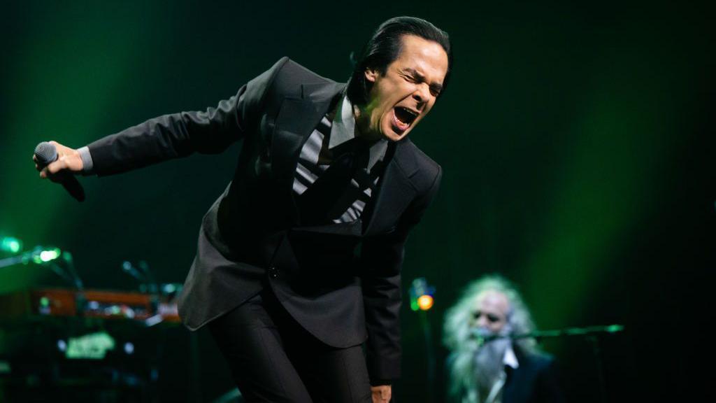 Cave sings in a dark suit with hair slicked back. He is holding a microphone in his outstretched arm.