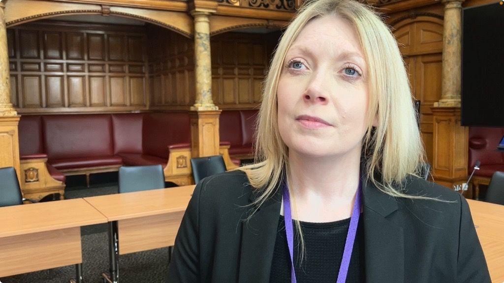 Head and shoulders shot of Natalie Hoy. She is wearing a black suit jacket and a purple lanyard around her neck.