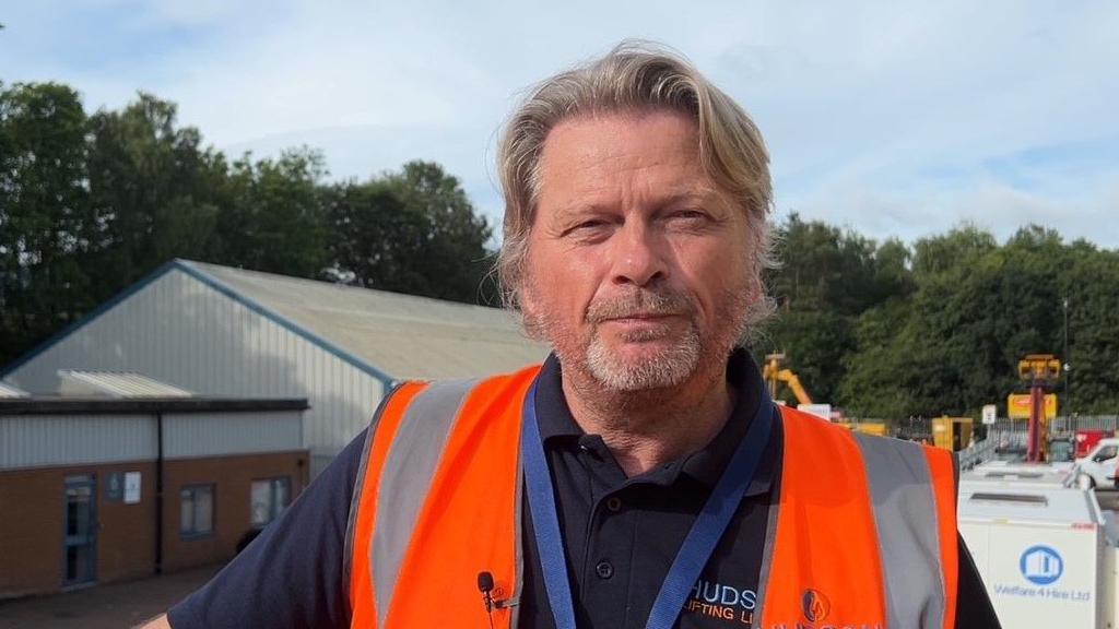 Managing director of Hudson Lifting, Jeff Hudson, has greying blonde hair and a grey beard and is wearing an orange high visibility jacket. He is on an industrial site with heavy plant machinery in the background.
